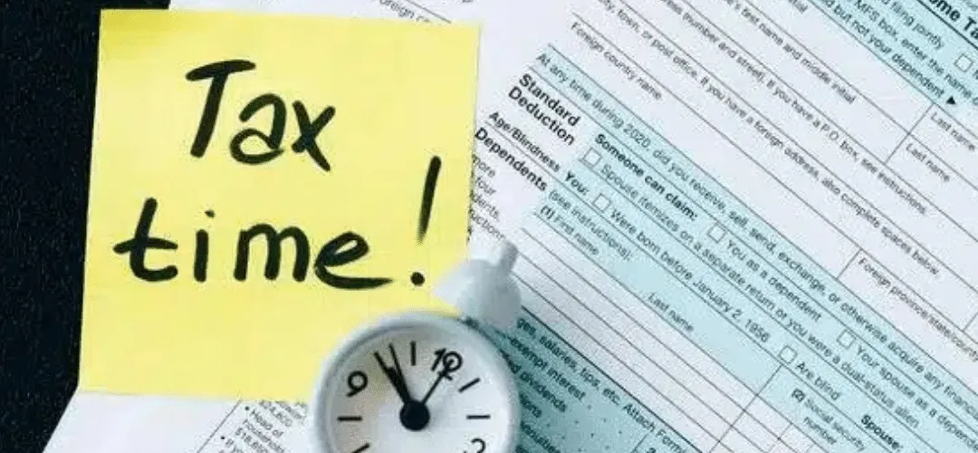 A yellow sticky note reading 'Tax time!' sits beside a small alarm clock on top of IRS tax forms, suggesting an approaching filing deadline.