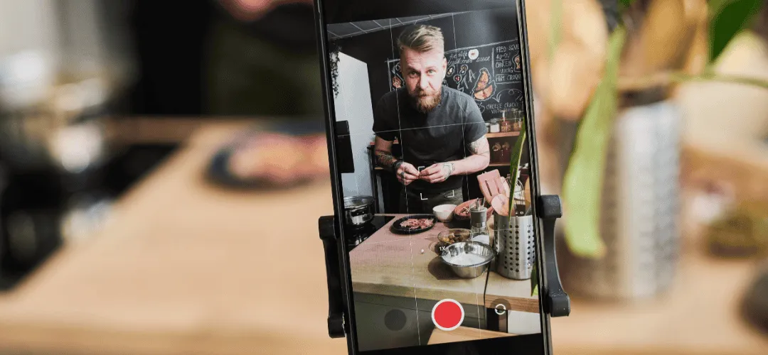 A smartphone mounted on a clip holder records a bearded man preparing food at a kitchen counter, with the red record button visible on the screen.