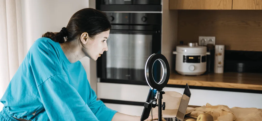 A woman in a teal top leans toward a ring light and phone setup on a small tripod while working on a laptop, filming content in a home kitchen setting.
