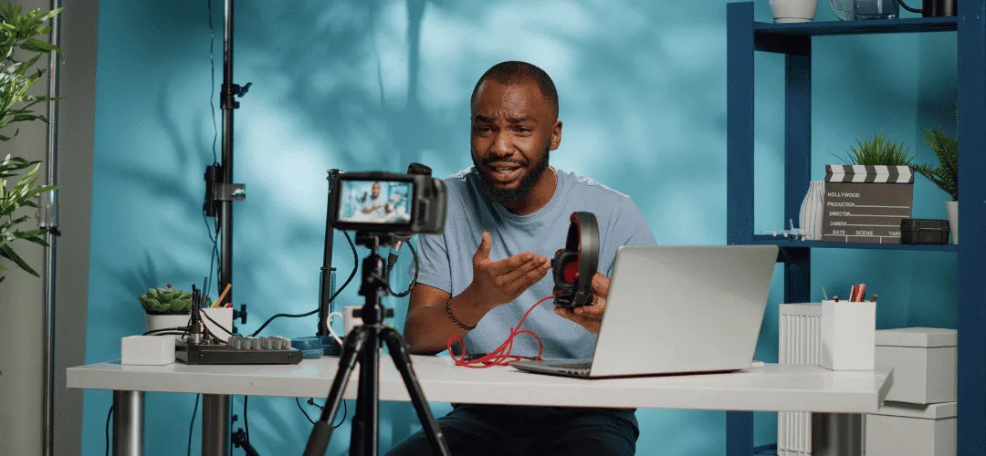 A man holds up a pair of headphones and gestures while recording a tech review video, with a DSLR camera on a tripod, laptop, studio light, and clapperboard visible in his blue-walled home studio.