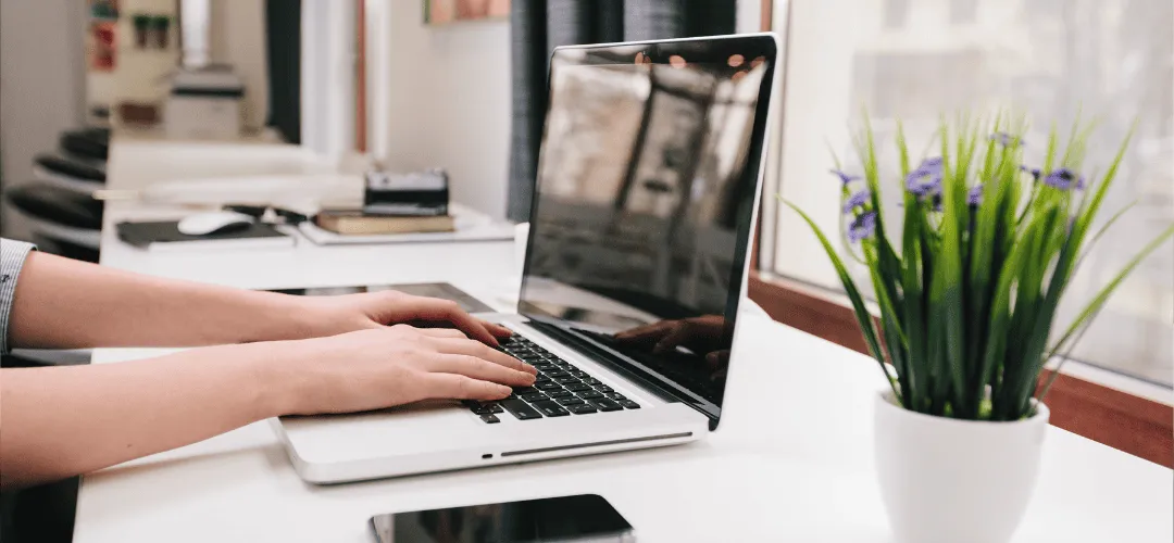 Close-up of hands typing on a laptop on a clean white desk, with a small potted plant with purple flowers beside it and a blurred home office in the background.