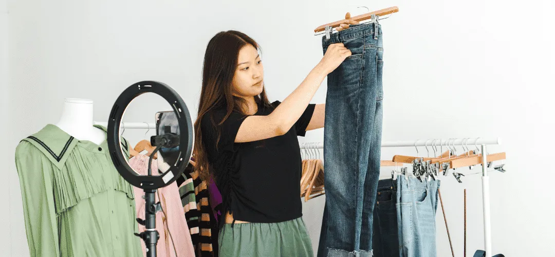 A WhatNot creator holds up a pair of jeans on a hanger while live-selling in front of a ring light with a phone, surrounded by clothing racks and a mannequin in a clean white studio.