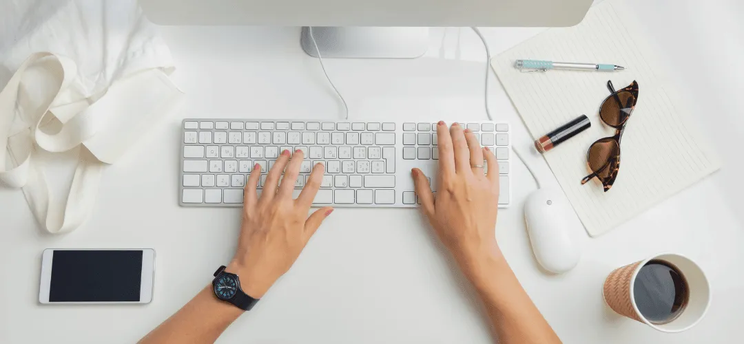 Overhead view of a woman's hands typing on an Apple keyboard, filing her taxes with sunglasses, lipstick, a pen, phone, mouse, and coffee cup arranged on a clean white desk.