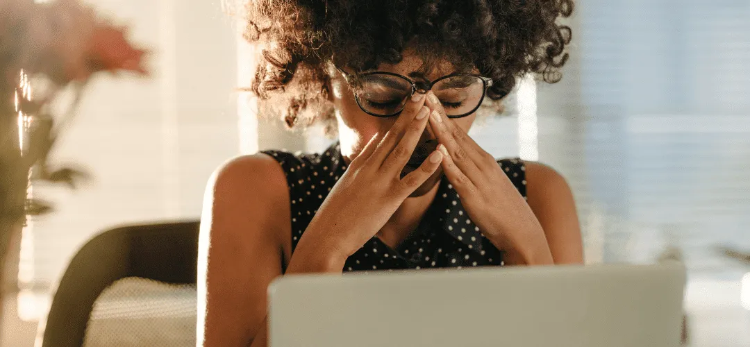 A woman in glasses and a polka-dot top presses her hands to her face in exhaustion or stress while filing past tax returns while sitting in front of a laptop in a warmly lit room.