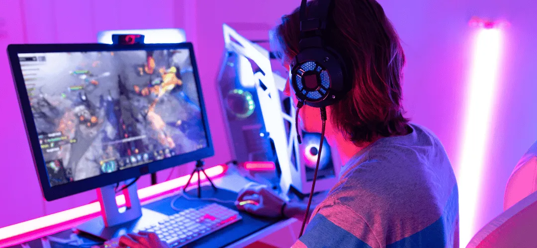 A person wearing large gaming headphones plays an action game on a monitor at an RGB-lit gaming desk with a white PC tower, bathed in pink and purple neon light.