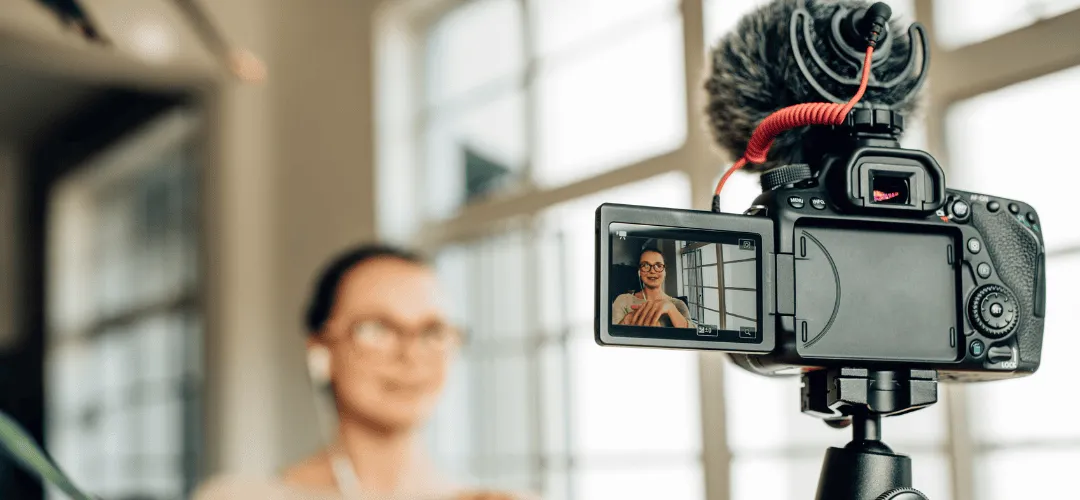 A DSLR camera with a shotgun microphone on a tripod shows a woman in glasses on its flip screen as she records herself in a bright, windowed room.
