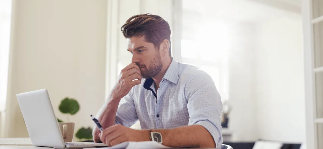 A worried-looking man in a plaid shirt holds a pen and rests his fist against his mouth, worrying about his tax audits, staring at documents beside a laptop in a bright, airy home office.