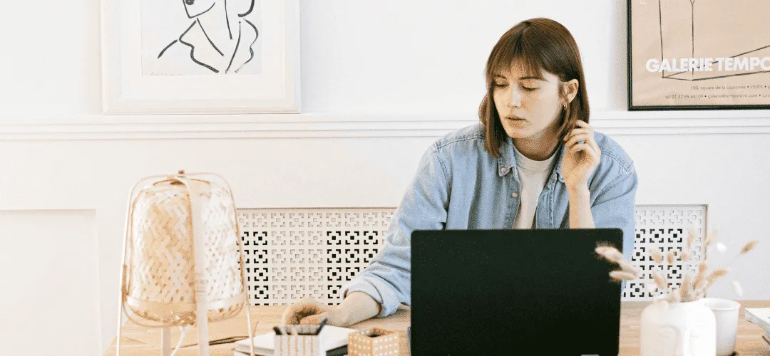A focused young woman in a denim shirt works at a laptop in a bright, minimalist home office decorated with framed art prints and a woven lamp.
