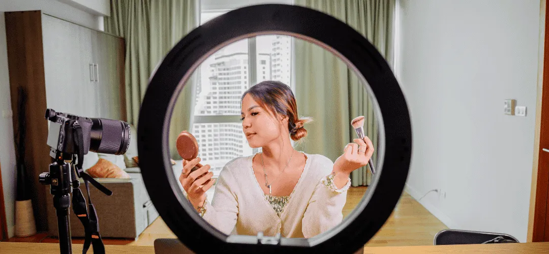 A woman holds a compact mirror and makeup brush while filming a beauty tutorial, framed through a large ring light with a DSLR camera on a tripod beside her in a bright apartment.