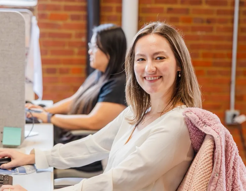 A woman smiling at her desk with colleagues working nearby