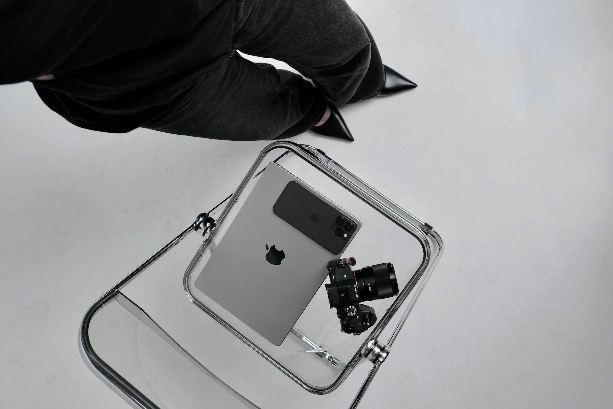 Overhead view of a tablet, phone and camera on a clear acrylic chair beside someone in black jeans and pointed shoes