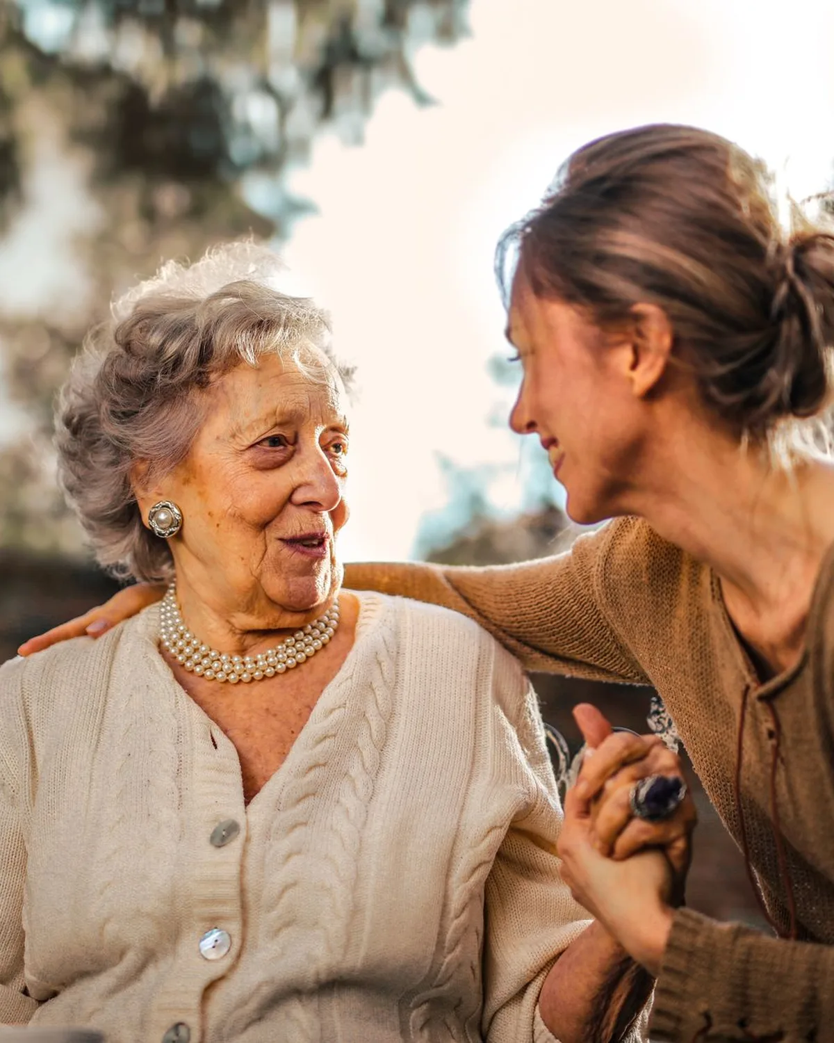 An adult daughter and her mother hold hands and smile in the garden.