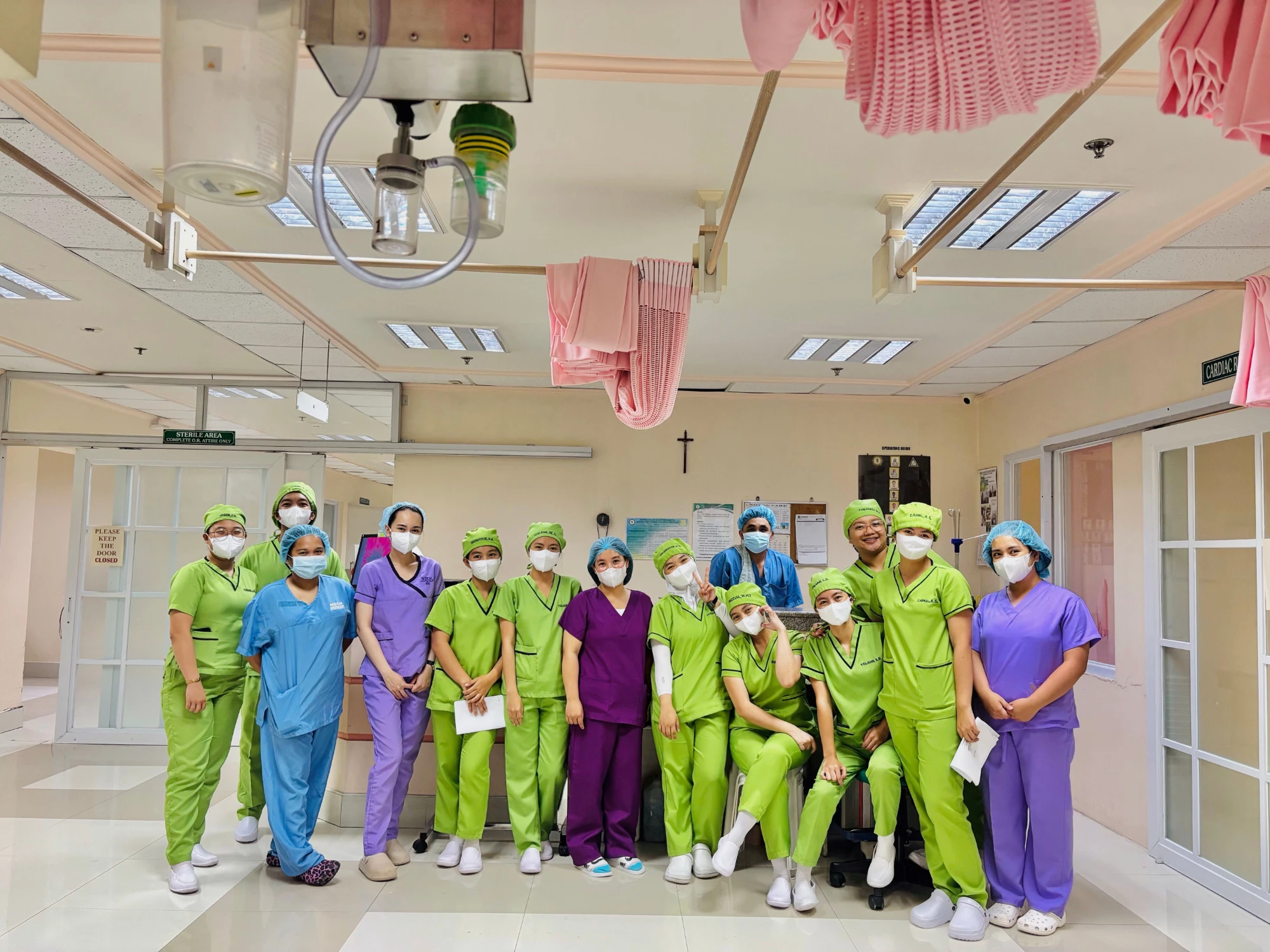 A group of nurses and surgical staff in colourful scrubs, caps and masks pose together in a hospital corridor outside the operating rooms.