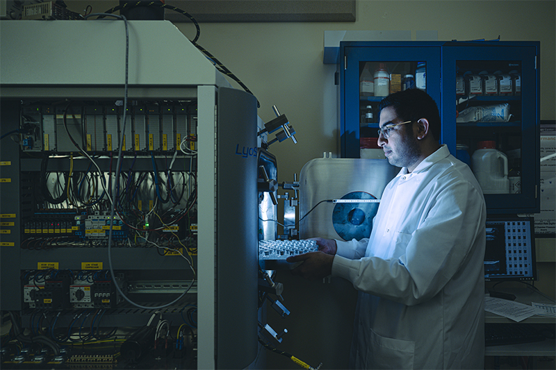 Researcher holding a tray of vials beside a lab-scale freeze dryer in a dimly lit lab
