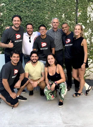 Group of eight smiling people posing outdoors in front of a green leafy wall, some wearing Rocket.Chat t-shirts.