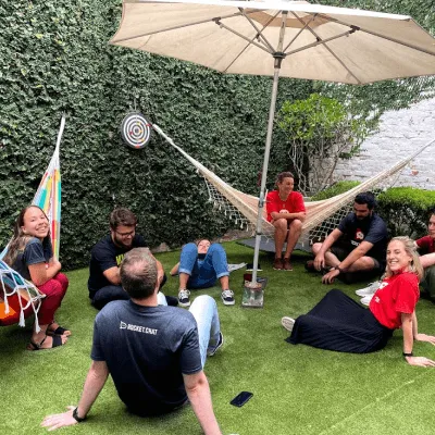 Group of seven people sitting and lounging on artificial grass and a hammock under a large patio umbrella in a backyard with green ivy on walls.