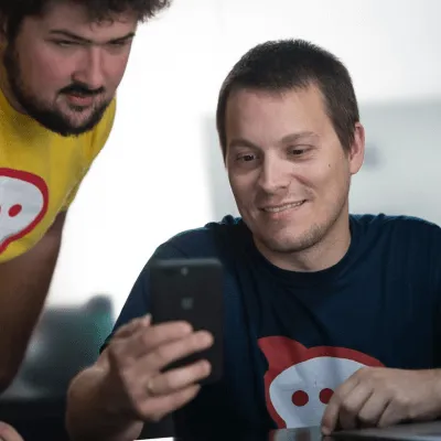 Two men looking at a smartphone together, one seated and smiling, the other standing and watching closely.