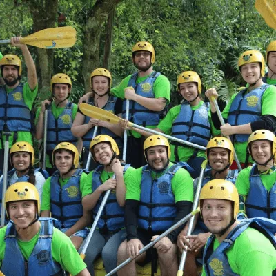 Group of people in green shirts, blue life jackets, and yellow helmets holding paddles ready for rafting in a lush outdoor setting.
