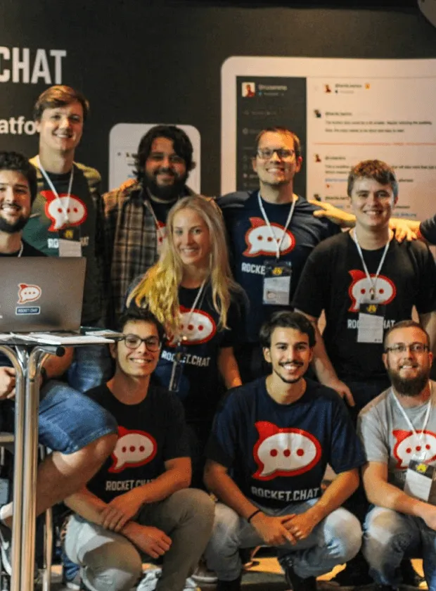Group of smiling people wearing Rocket.Chat t-shirts posing together indoors with a laptop showing the Rocket.Chat logo.