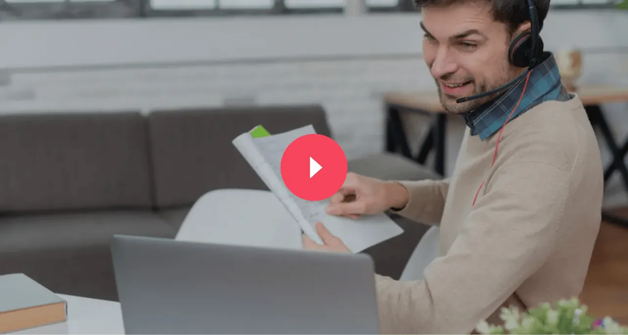 Man wearing headset, smiling and pointing at documents while sitting at a desk with a laptop.