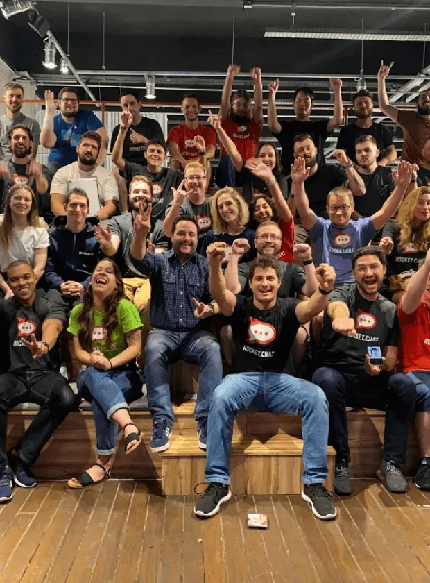 Large group of people seated on wooden steps indoors, smiling and cheering, many wearing Rocket.Chat t-shirts.