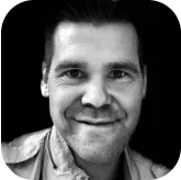 Close-up black-and-white portrait of a smiling man with short dark hair against a black background.