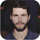Man with dark hair and beard wearing a dark blue shirt against a dark background.