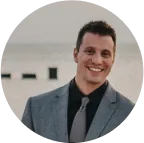 Smiling man in a gray suit and black shirt with a dark tie standing outdoors.