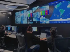 Three people in a control room monitoring multiple large screens displaying maps and satellite images.