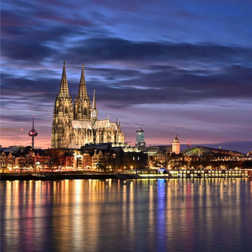 Evening view of Cologne Cathedral and city skyline with reflections on the Rhine River.