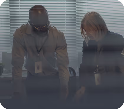 Two people wearing lanyards standing and leaning over a table in a dimly lit room with blinds.