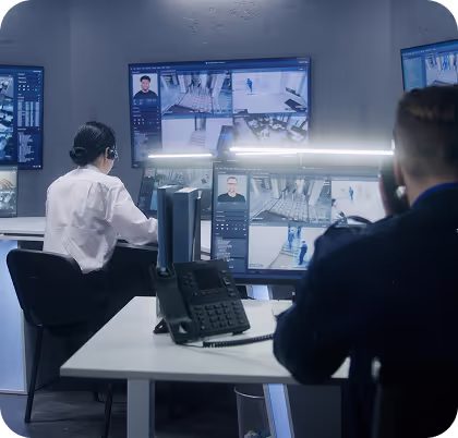 Two security personnel monitor multiple surveillance camera feeds on large screens in a control room.