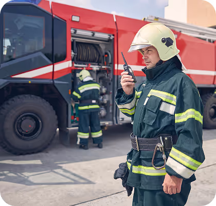 Firefighter in protective gear using a radio near a red fire truck with another firefighter accessing equipment compartments.