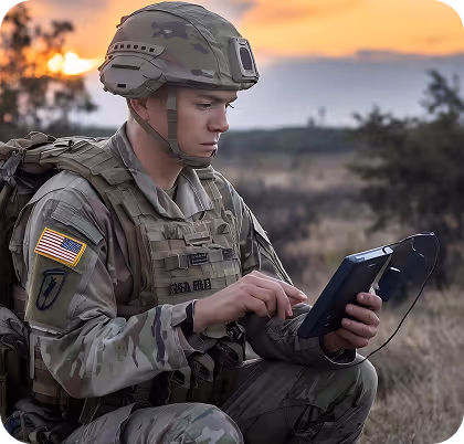 Soldier in camouflage uniform and helmet using a handheld device outdoors at sunset.