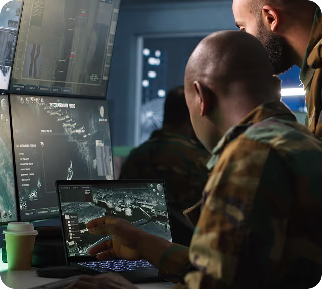 Two men in military uniforms analyzing maps and data on multiple computer screens in a dimly lit room.