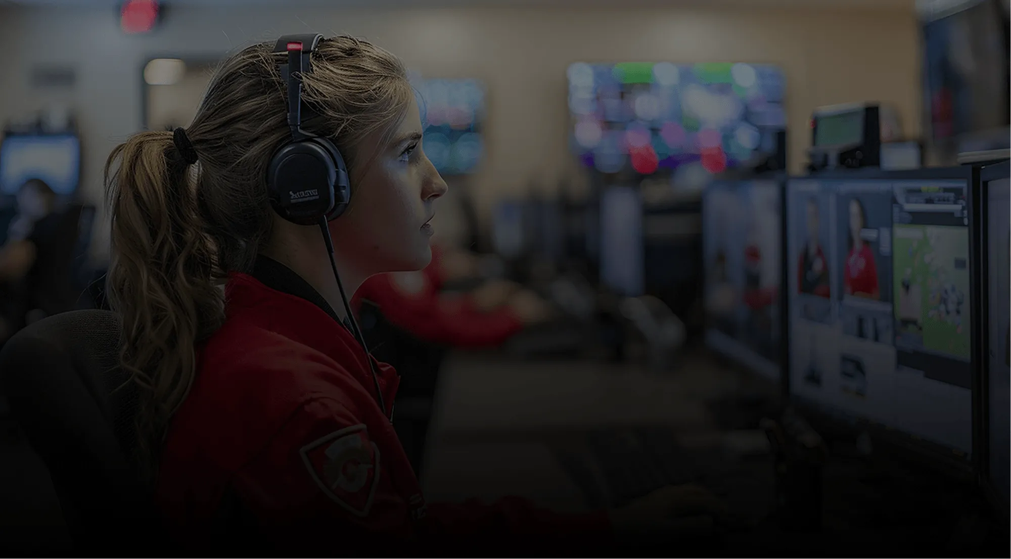 Woman wearing a headset and red uniform working at a computer with multiple monitors in an office environment.
