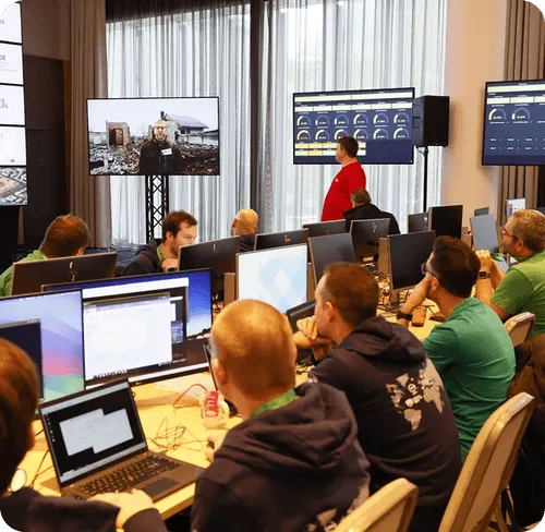 Group of people seated at desks with multiple computer monitors watching a presentation on large screens in a conference room.