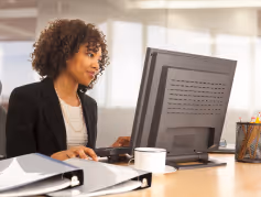 Woman with curly hair working on a desktop computer at an office desk with binders and a coffee cup.