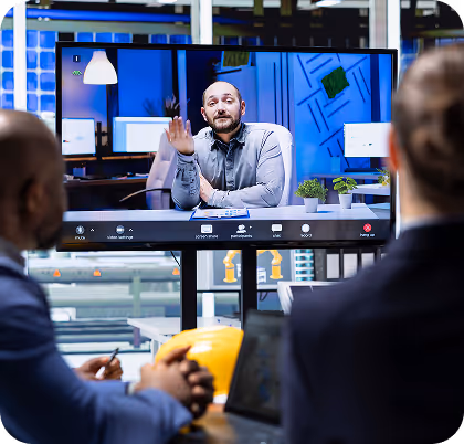 Two people attending a video conference call with a man speaking on a large screen in a modern office setting.
