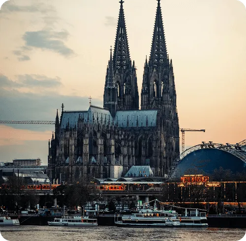 Cologne Cathedral with twin spires at sunset, riverfront with boats, and a nearby illuminated sign reading 'RheinEnergie'.