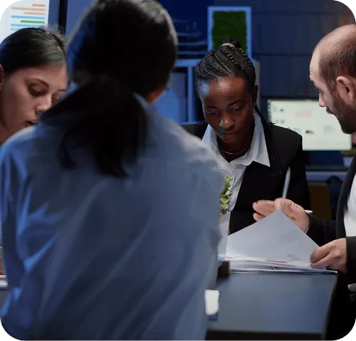 Four professionals reviewing documents collaboratively in an office setting at a table with computer screens in the background.