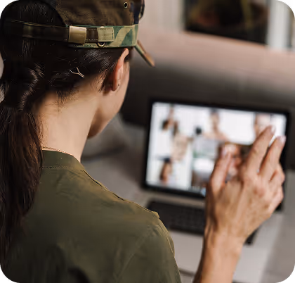 Person in military-style clothing and camouflage cap waving at a laptop screen during a video call.