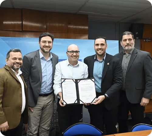 Five men standing indoors, two in center holding and displaying a signed document, all smiling.
