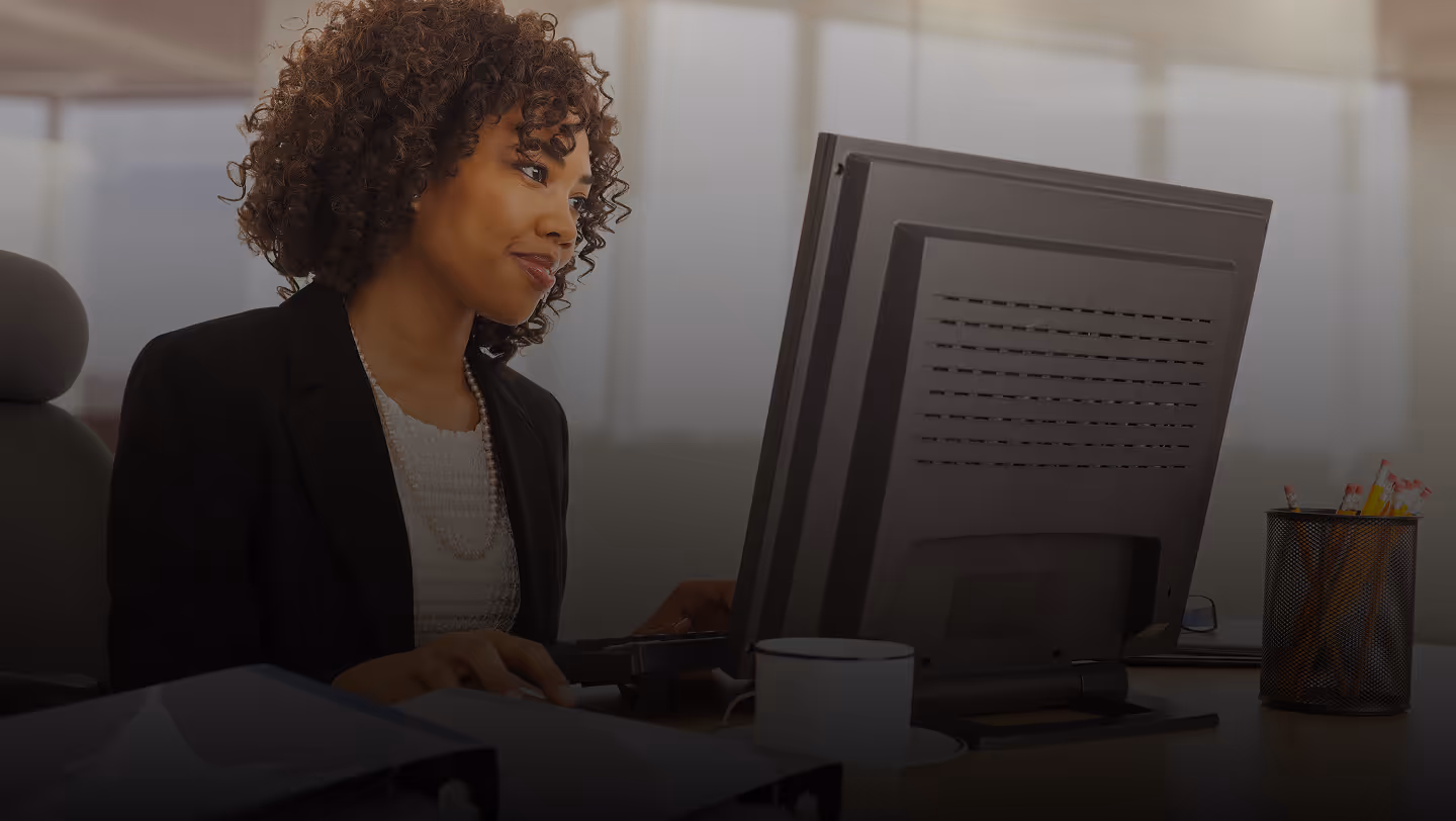 Woman with curly hair working on a desktop computer in a modern office setting.