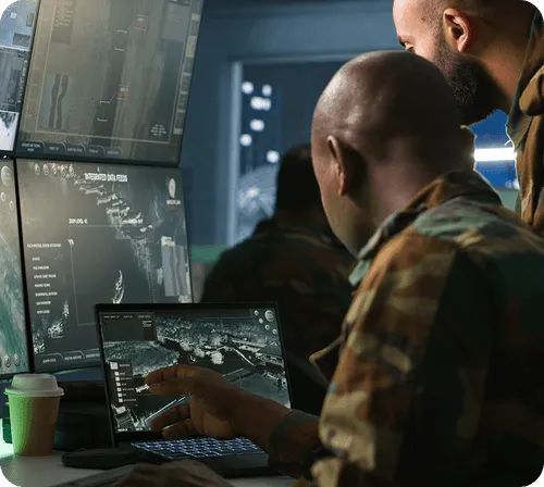 Two military personnel in camouflage uniforms analyzing tactical maps and data on multiple screens and a laptop in a dark command center.