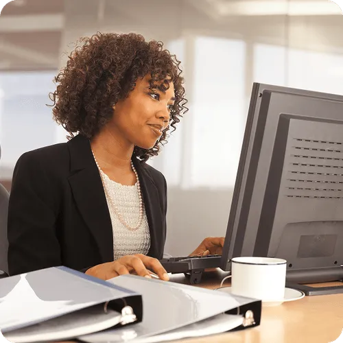 Young woman with curly hair working at a desktop computer in an office, with binders and a coffee cup on the desk.