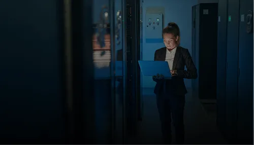 Woman in a dark server room holding and looking at an open laptop.