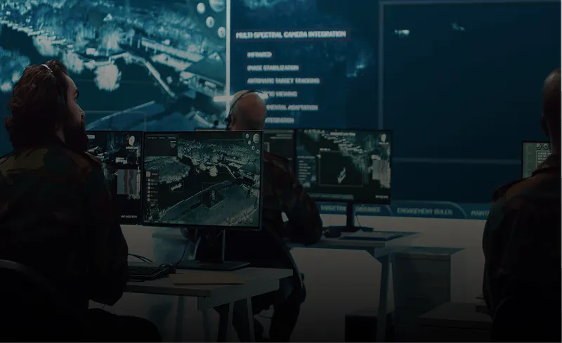 Three people sitting at desks with multiple computer monitors displaying detailed aerial maps in a dark command center.