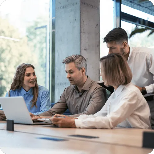 Four colleagues collaborating around a laptop in a modern office with large windows.