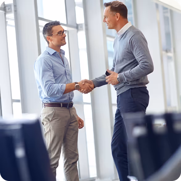 Two men shaking hands and smiling inside a modern office with large windows.